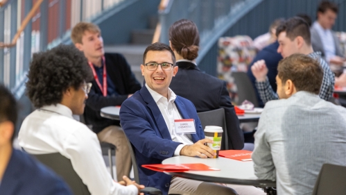 A man in a blue jacket is sitting at a table and smiling while engaging with colleagues.