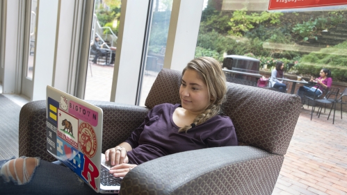 A female student reclines in a student lounge while typing on a laptop..