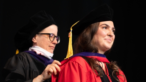 woman in black robe and cap hooding a woman in red robe and black cap on stage