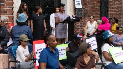 people protesting with signs and a woman speaking behind a podium