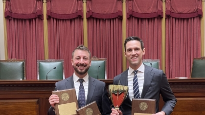Two men in business suits smiling and holding plaques and a trophy in courtroom