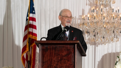 man wearing glasses speaking at lectern with American flag behind him