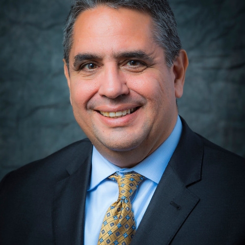 Man smiling wearing blue shirt, yellow tie, and black suit jacket