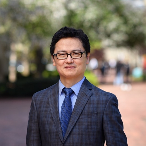 Man grinning with glasses wearing a blue shirt, blue tie, and plaid suit jacket