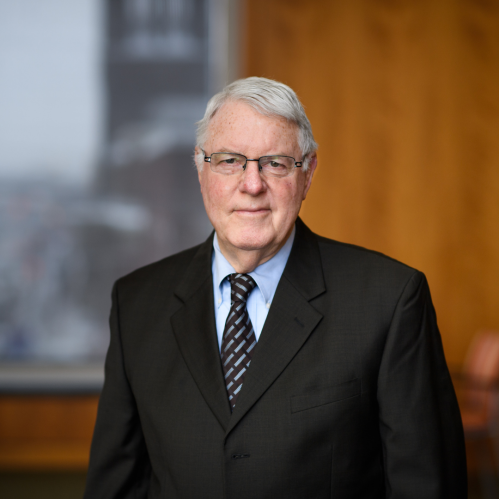 Man grinning wearing glasses, a blue button down shirt, a grey and black tie and a suit.