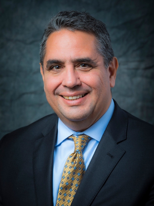 Man smiling wearing blue shirt, yellow tie, and black suit jacket