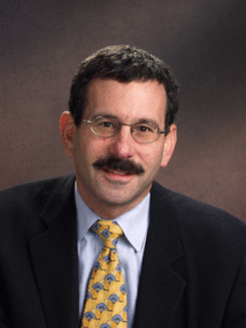 Man grinning wearing glasses, blue shirt, yellow tie, and suit jacket