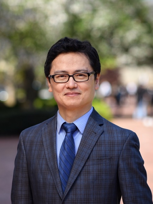 Man grinning with glasses wearing a blue shirt, blue tie, and plaid suit jacket