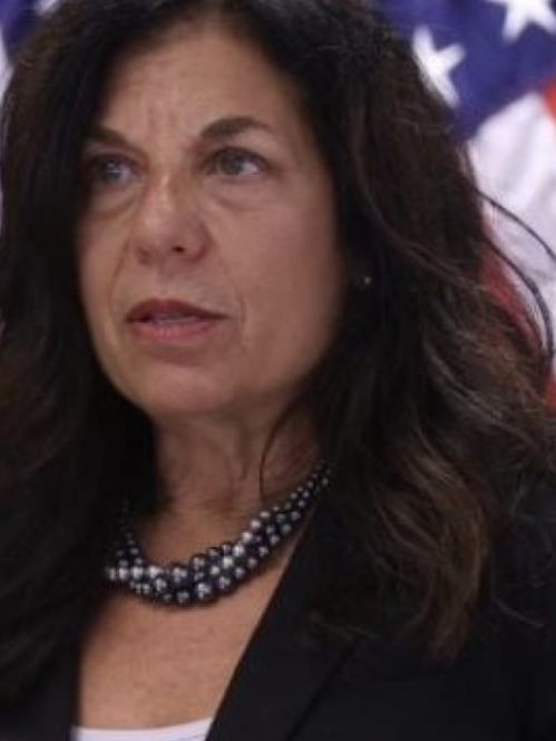 Woman grinning in front of an American flag wearing a black blazer.