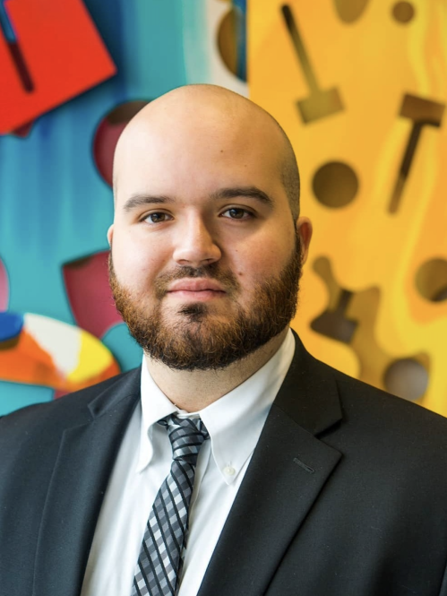 Man grinning wearing white button down and grey suit jacket in front of colorful background