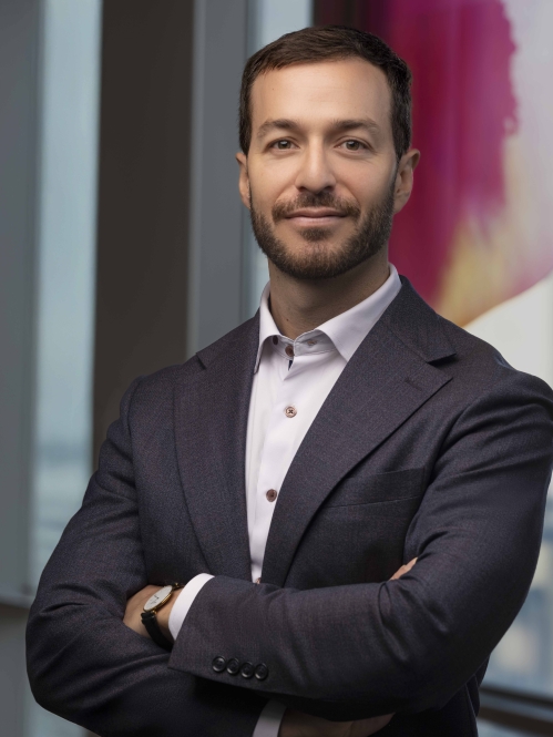 man in dark grey suit with arms folded in front of large window