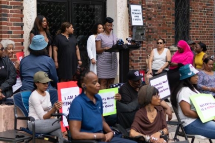 people protesting with signs and a woman speaking behind a podium