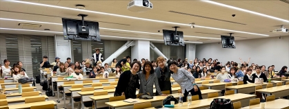 Four people standing and smiling in front of large class with students seated