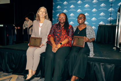 three women seating on edge of stage smiling with two of them holding plaques