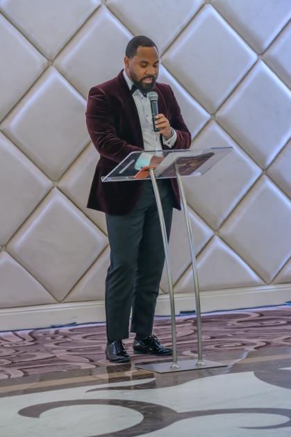 man in formal wear speaking at clear podium in front of quilted wall