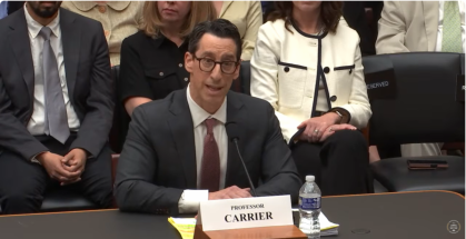 man in business suit seated at table and speaking into mic with name card in front of him