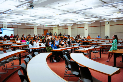 large classroom with people seated at semi circular tables and woman speaking at front of class