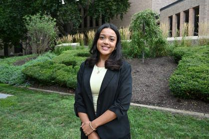 woman in black blazer and light green shirt smiling outdoors with hands folded in front of her