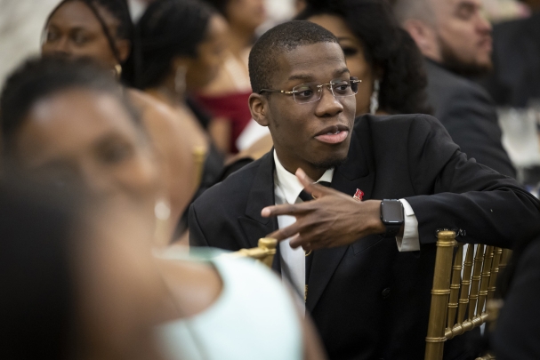 Man in black suit and glasses seated at table in large dining hall