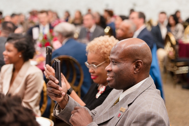 man and woman holding phones up and smiling seated at table in large dining hall