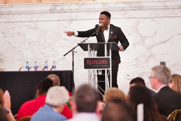 man in black suit and bow tie speaking and pointing on stage at podium