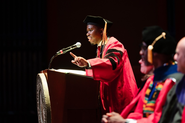 man speaking at podium on stage wearing red graduation robe and black cap