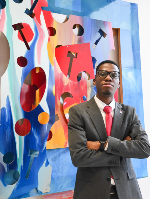 man in gray suit with folded arms and glasses standing in front of colorful mural