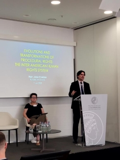 man in a suit standing behind a podium and a woman sitting next to him
