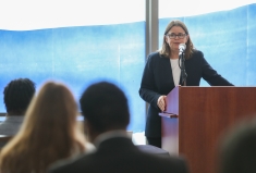 woman in black blazer and white shirt speaking at brown podium in front of blue and white window