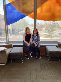 two women seated on bench side by side and smiling in front of colorful window