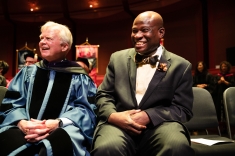 Man in blue graduation robe seated next to man in suit on stage