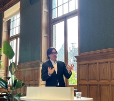 man in black suit and white shirt with classes speaking in front of window