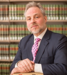 man in suit resting folded hands on top of book in front of large bookcase with green books