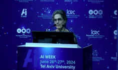 woman wearing glasses in speaking at podium surrounding by white logos on blue background