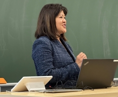 Woman speaking in dark blue suit in front of chalkboard and laptop in front of classroom